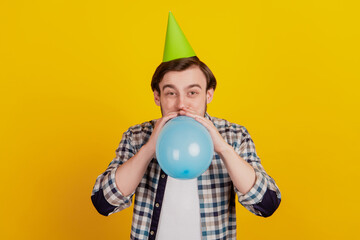 Photo of young guy blow balloon wear paper cone hat birthday party decoration isolated yellow color...