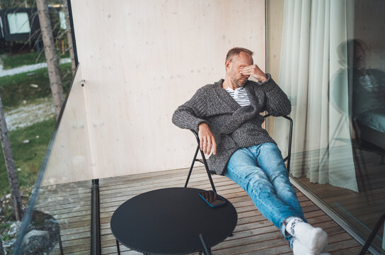 Portrait of a tired Middle-aged man dressed in open cardigan, jeans, and warm socks sitting on forest house balcony. He massaging tired eyes after. Vision health problems concept image.