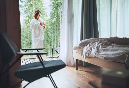 Woman dressed white bathrobe standing on forest house balcony and enjoying fresh air and morning teacup. Inside a Scandinavian interior design room view with a not made bed. Cozy living concept image.