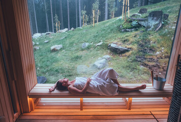 Relaxing Woman wrapped white towel lying on the wooden bench in Hot Finnish sauna with a huge wide...
