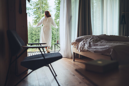 Woman dressed white bathrobe standing on forest house balcony and enjoying fresh air with nature view. Inside the Scandinavian interior design room with a not made bed. Living in wild concept image.