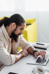 serious hispanic man using laptop near blurred notebooks on desk