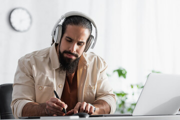 hispanic designer in headphones drawing with stylus near blurred laptop
