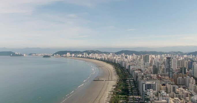 beautiful beach with city buildings on the shoreline. drone flying over buildings and shore. slow side camera movement with mountains in the background. Santos. Blue sky