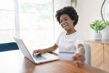 Smiling young African female entrepreneur working online with a laptop while sitting at her kitchen...