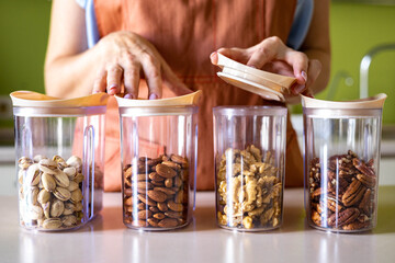 Closeup woman hands in apron placing different nuts into glass jar storage container at kitchen