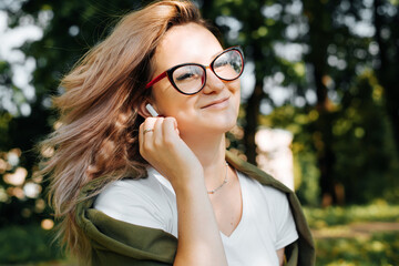 Portrait of smiling young woman listening audio on wireless portable earphones, teenage girl in red glasses enjoying music outdoors. Selective focus on trendy white headphones