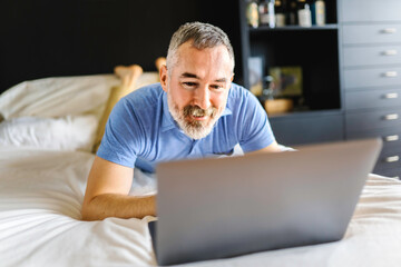 nice mature man at hotel with grey beard relaxing on bed with laptop