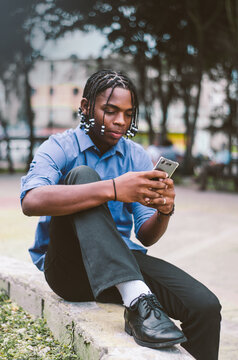 young latino dominican man with dark skin and curly and braided hair in the city park in a blue shirt, sitting sending a message with his smartphone
