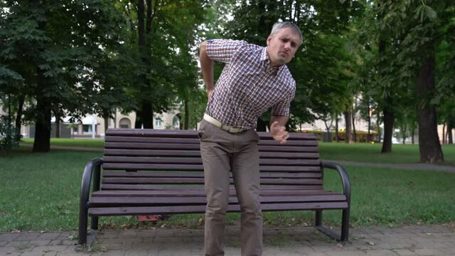 Mature man with grey hair wearing shirt stands up from bench to greet friend and suddenly feels severe back pain in city park