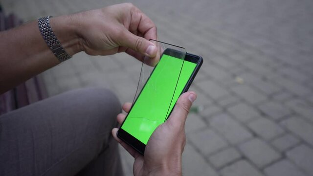 Man in trousers with wristwatch removes cracked protective glass from smartphone green screen and cleans with fingers closeup