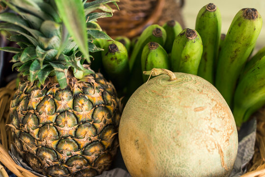 A pineapple, melon and green Lacatan banana in a basket, on display at a small fruit stand.