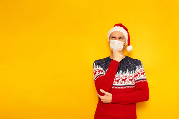 man in New Year sweater with reindeer, medical mask, Santa Claus hat holds his hand under his chin in thoughtful pose thinking about choice gifts. isolated yellow background with space for text