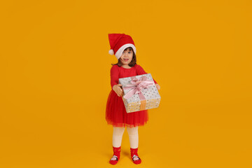 a little beautiful girl in a red Santa Claus hat dress holds a gift box for the new year in her hands