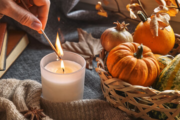 Hand with burning match lighting a candle on the windowsill with cozy autumn still life with pumpkins, knitted woolen sweater and books. Autumn home decor. Cozy fall mood. Thanksgiving. Halloween.