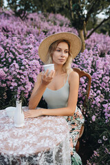Fototapeta premium Young blonde woman sitting at the table with cup of tea in flower garden