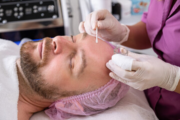 Cosmetologist applies a medical on male face. Adult man in a spa salon on cosmetic procedures for facial care. White middle-aged man getting beauty treatment therapy.