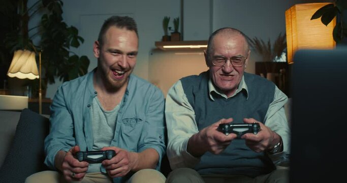 Happy senior 70s old man playing fun console videogame using joysticks bonding with young adult son at home on couch.