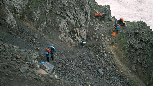 Climbers take turns walking one after another in the mountains holding on to the rope. A group of tourists with backpacks with equipment on the rocks are ferried along the railing