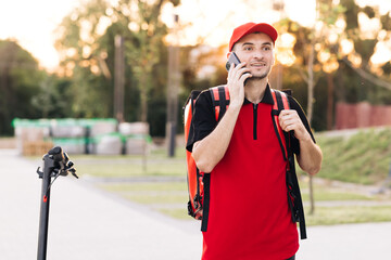 Male courier with isothermal food case box arrives on a electric scooter to the entrance to the house and calls for client. Food delivery guy with red backpack searching delivery addresses
