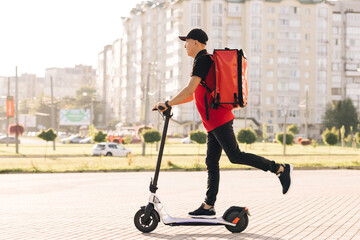 Man courier food delivery with red thermal backpack rides the street on an electric scooter deliver online order client customer. Deliveryman worker employee in red uniform. Excellent delivery