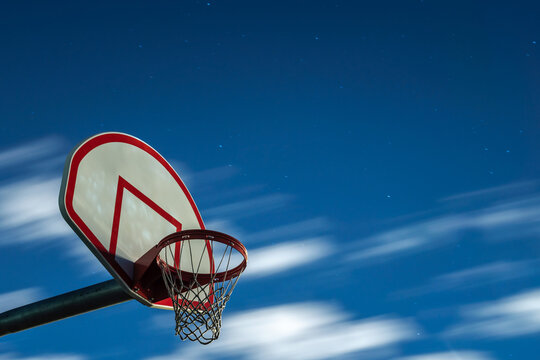 A long exposure photograph of a dirty orange and white playground basketball hoop, rim and net with motion blurred clouds in a dark blue night sky in the background.