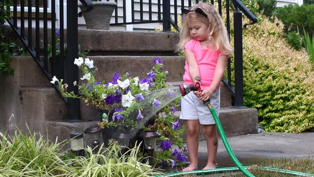 Toddler girl watering the garden on a summer day. Child playing with the water hose. Toddler summer fun activities