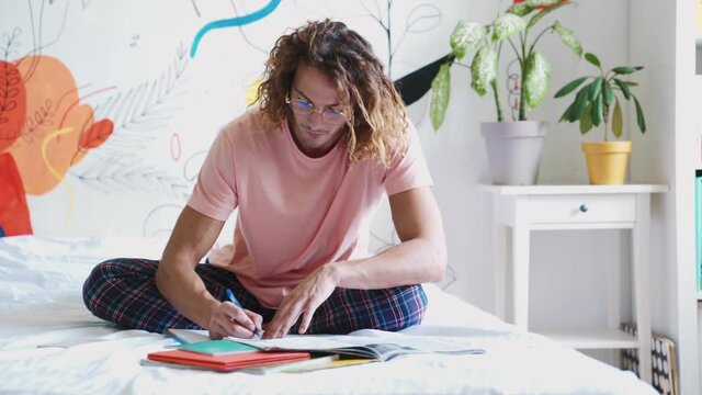 Concentrated long-haired man doing homework while sitting on bed