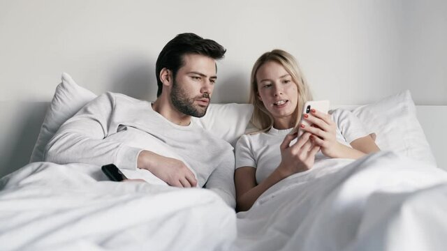Confident woman showing her boyfriend something on the phone and talking while lying on the bed