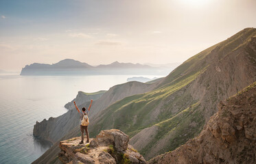 Young man travels alone on the backdrop of the mountains standing on top of a mountain with raised hands, the lifestyle concept of traveling outdoors.