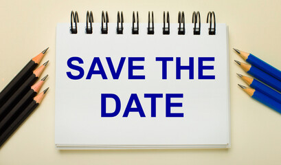 On a light background, a white notebook with the text SAVE THE DATE and black and blue pencils on the sides.