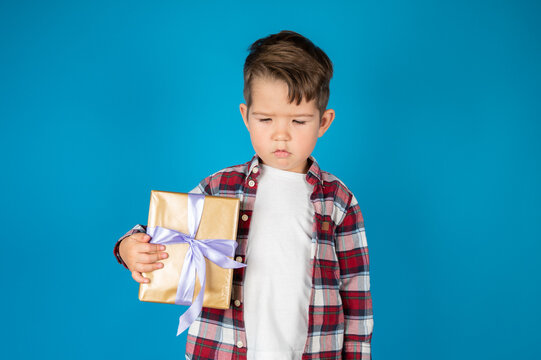 Sad Little Boy In Plaid Shirt With Gift Box On Blue Background. Child Displeased With Bad Present.