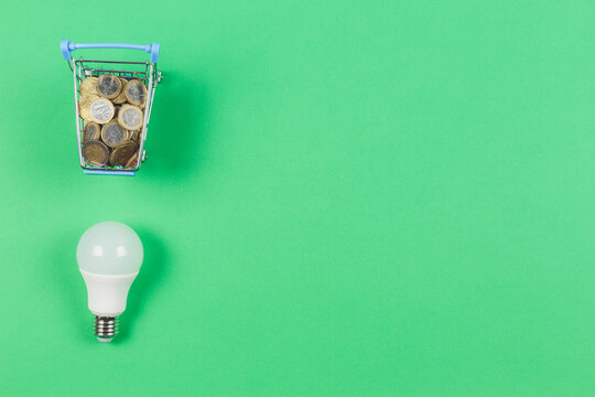 Top view to mini shopping trolley with euro coints and light lamp bulb on light geen background. Electricity cost and expensive energy concept