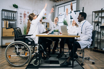Diverse group of qualified doctors smiling cheerful and gesturing with arms raised while sitting at...