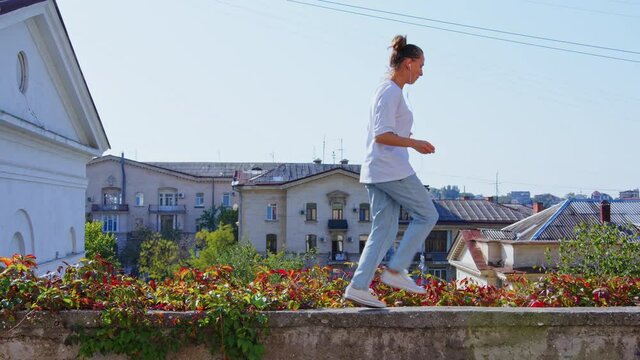 Girl in jeans performing modern dances on retaining wall near flowerbed. Cozy town with small houses, roofs. Female dancer dancing hip hop, shuffle, contemporary. Woman rhythmically moving at street