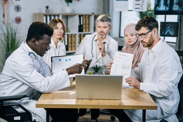 Fototapeta premium Five multi ethnic medical specialists showing various charts and graphs during video call on modern laptop. Group of competent doctors having online conference at office.