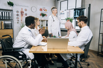 African american doctor who using wheelchair listening to male lecturer with his international colleagues during seminar. Aged caucasian therapist talking about x ray scan at boardroom.