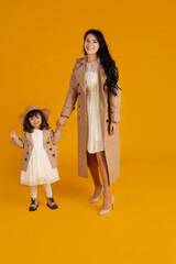 a little beautiful girl in a white dress beige raincoat and beige hat beige shoes is standing next to her mother in a beige trench coat on a yellow background