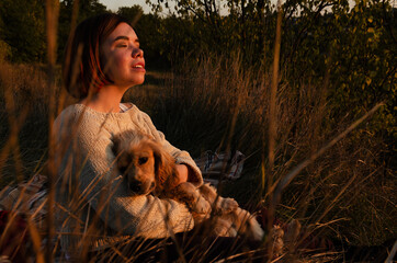 Thoughtful woman with short hair sitting on the ground during golden hour with her pet dog, enjoying sunset.