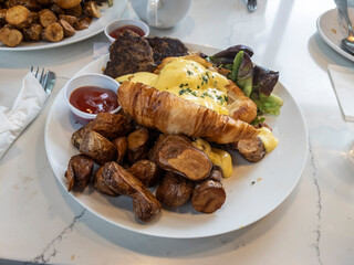 Top down view of a large breakfast spread of eggs benedict, fingerling potatoes, and turkey sausage patties on a marble table in an eatery
