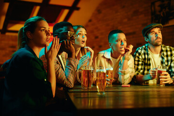 Multi-ethnic group of friends watch sports match with anticipation while drinking beer in pub.