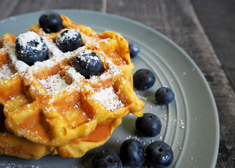 Belgian gluten-free pumpkin waffle, sprinkled with powdered sugar and blueberries, lies on a gray plate on a gray table, side view. vegan breakfast