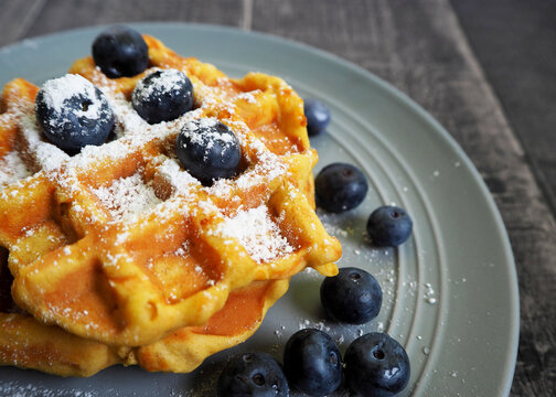 Belgian gluten-free pumpkin waffle, sprinkled with powdered sugar and blueberries, lies on a gray plate on a gray table, side view. vegan breakfast