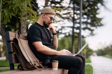 Thoughtful guy with laptop doing remote work outside while sits on bench in city park. Distance Job 