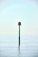 An isolated marker post off the beach in Eastbourne, East Sussex. Light grey cloud and calm sea.