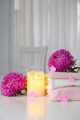 Set of fresh towels of pink and white color with purple aster flowers, candle and pink hearts on white table. Spa or massage salon concept. Romantic surprise for Valentine`s Day. Copy space.