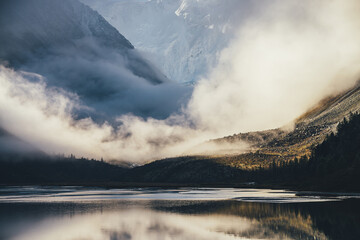 Mountain landscape with shiny lake and silhouettes of coniferous trees on dark rocks with view to high snowy mountain wall in low clouds in golden sunshine. Layered scenery with shadow and sunlight.