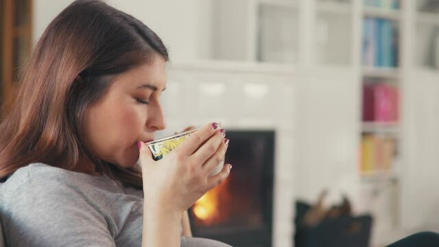 Beautiful pregnant woman with brown hair stitting in front of cosy fireplace drinking tea shot in 4k