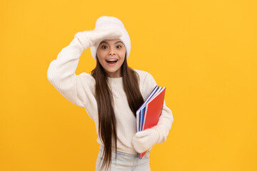 amazed teen girl ion winter hat and gloves hold notebook on yellow background, school fun