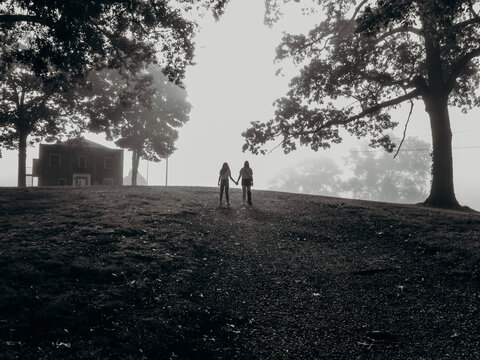 Black and white photo of two girls in silhouette holding hands between tall trees with fog